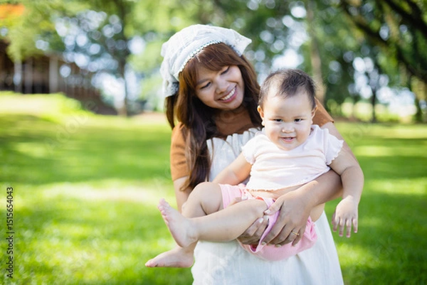 Obraz Family enjoys leisure time outdoors with picnic concept as woman happily holds baby in green park filled with trees and sunlight creating joyful moment for people