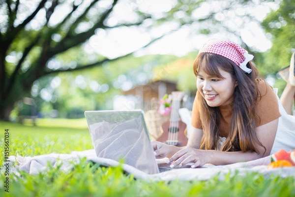 Obraz Young woman enjoys leisure time on picnic blanket using laptop in park surrounded by green grass and trees, capturing concept of people relaxing outdoors with family