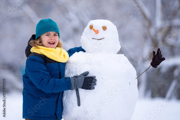 Fototapeta Kid play with snowman in winter. Snowman with a carrot nose on a winters day. Child making snowman. Little boy building snowball for funny snowman in snowy winter park. Snowmen in a snowy winter park.