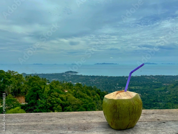Fototapeta Coconut with a drinking straw on a wooden table with a view of Samui island from the terrace of the house on Lipa Noi Hill, Koh Samui