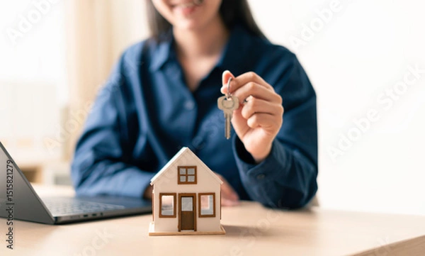 Fototapeta woman holding house keys above a miniature home model next to a laptop in a home office to represent property purchase completion