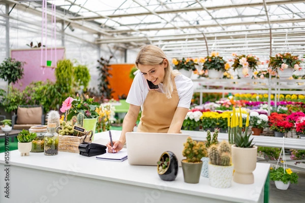 Obraz A Caucasian woman works at a florist shop, taking orders over the phone and using a laptop to manage sales while surrounded by various flowers and plants.