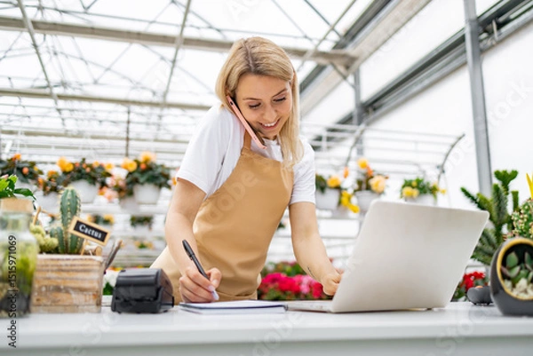 Obraz A Caucasian woman working in a florist shop, taking a call and using a laptop while writing.