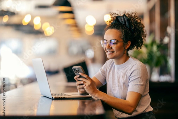 Fototapeta Cheerful interracial remote worker sitting in cafe with laptop in front of her and texting messages.