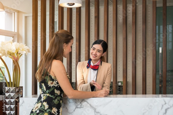 Fototapeta Friendly hotel staff in blazer with striped scarf warmly welcomes guest in sunflower dress at marble reception counter for room accommodation check in