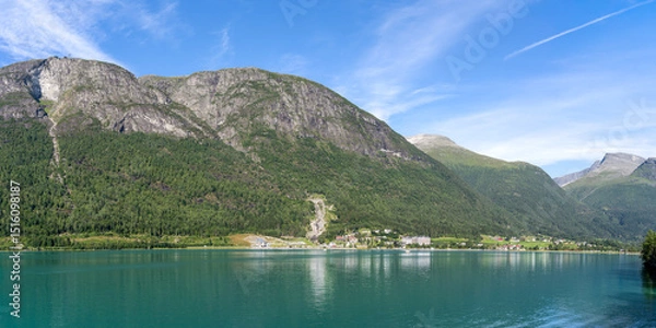 Fototapeta Panoramic view of Loen village, Norway, with the Innvikfjorden fjord in the foreground and majestic mountains and Hoven peak behind under a sunny sky, a typical northern fjord landscape