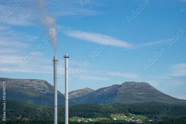 Fototapeta Two industrial chimneys release smoke into the clear blue sky, contrasting with the lush forest, green fields, and rocky mountains of the Norwegian landscape