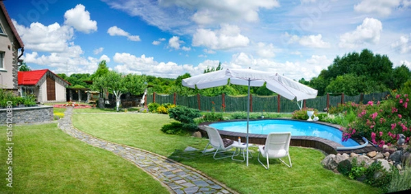 Obraz Beautiful landscape design of a country house with a well-groomed lawn, a swimming pool, an umbrella, chairs and a flowering bush. Lentious summer rest in nature.