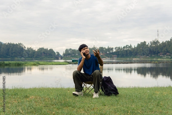 Fototapeta A young man sitting on a chair at lake with relaxing mood,  smiling and engaging in cheerful phone conversation.