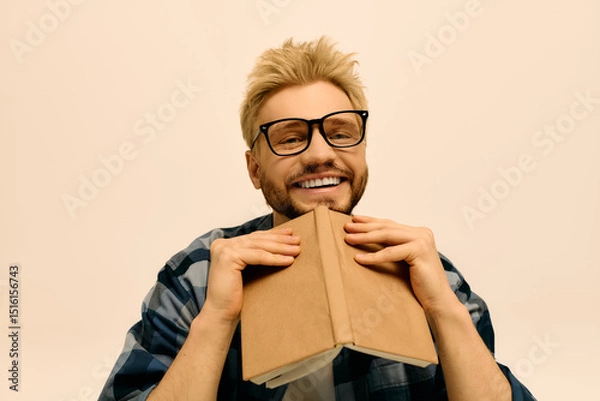 Fototapeta Handsome young man with glasses joyfully holding a book and smiling in a cozy indoor setting