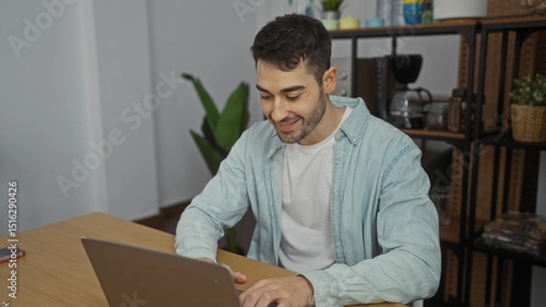Fototapeta Young hispanic man smiling while working indoors on laptop in office room with plants and wooden furniture, showcasing a productive and modern workplace setting