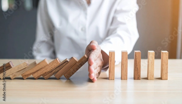 Fototapeta Person in white shirt stops falling wooden dominoes with their hand, symbolizing intervention, risk management, and prevention in business or personal context