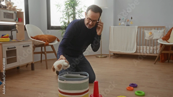 Fototapeta Man organizing toys in cozy bedroom while talking on phone, surrounded by colorful baby items in modern home environment.