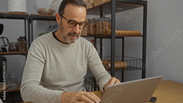 Fototapeta Mature man working on a laptop in an office environment, focusing intently amidst organized shelves, suggesting a professional workspace with a modern interior design.