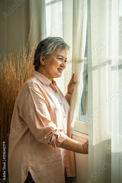 Fototapeta Smiling senior Asian woman looking out window with sunlight and positive mood