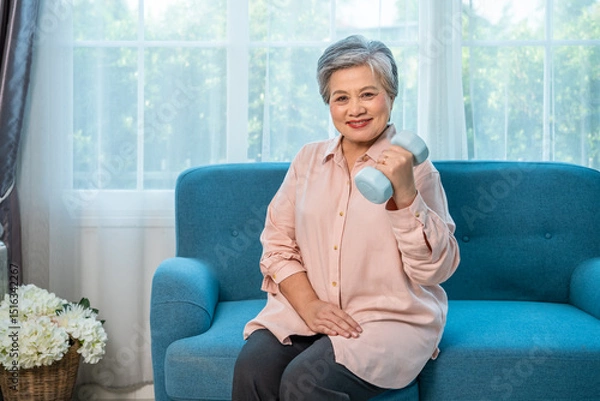 Fototapeta Senior Asian woman exercising at home with dumbbell for healthy aging and strength, looking at camera