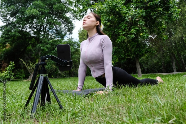 Fototapeta A woman is practicing yoga outdoors, following an online session on her phone. She sits on the grass in a peaceful natural setting, focused on the screen in front of her. 