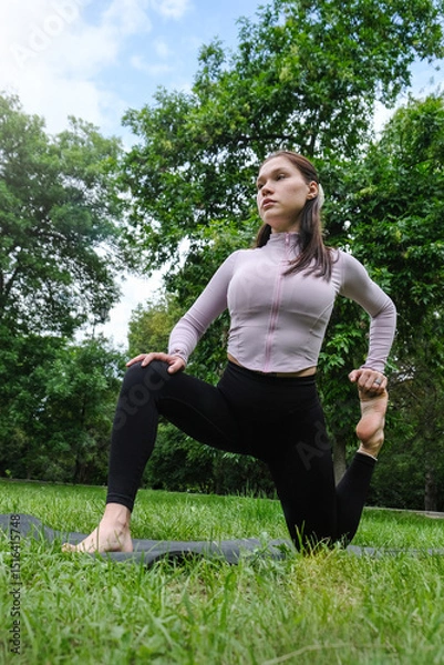 Fototapeta The photo shows a woman doing yoga in a natural setting, barefoot on the grass. Her body is steady in a graceful pose, and her face is calm and focused. 
