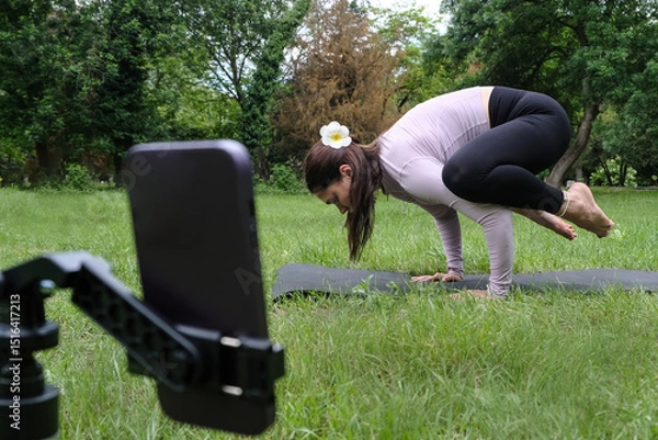 Fototapeta A woman is practicing yoga outdoors, following an online session on her phone. She sits on the grass in a peaceful natural setting, focused on the screen in front of her. 