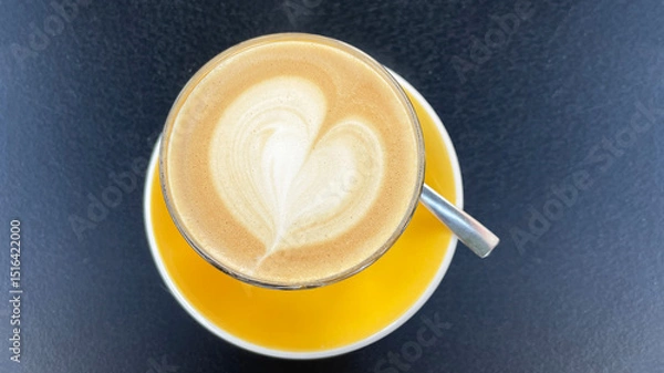 Fototapeta Cup of coffee with heart shape latte art on yellow saucer on dark cafe table
