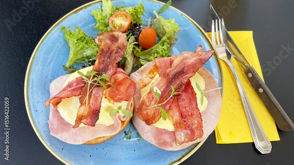 Fototapeta eggs benedict served with crispy bacon, ham and hollandaise sauce on brioche bun and side salad for breakfast or brunch