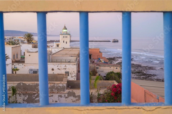 Fototapeta Sidi Ifni coastal landscape with lighthouse viewed through blue railing at sunset