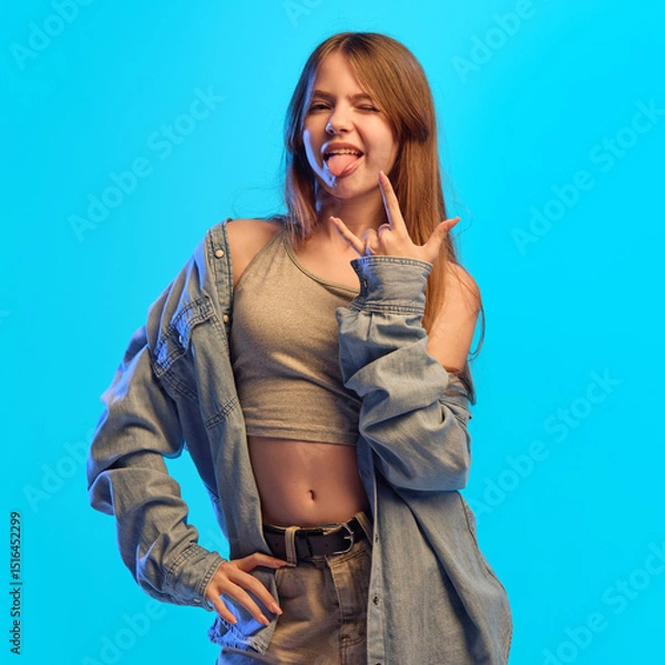 Fototapeta Teen girl sticks out tongue and makes rock gesture with fingers while posing in neon light against blue studio background showing bold attitude. Concept of playfulness, humor, self-expression
