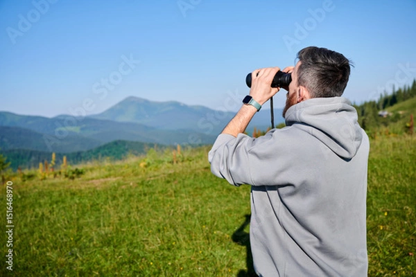 Fototapeta Man stands in grassy field, using binoculars to observe the distant mountains, wearing gray hoodie, smartwatch on his left wrist. Rolling green hills and clear blue sky, creating tranquil scene.