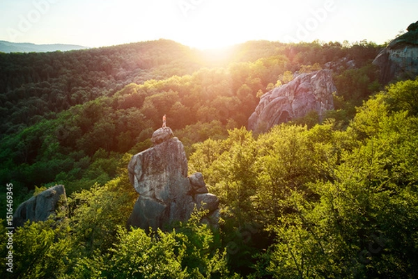 Fototapeta Aerial view of climber standing atop rock formation at Dovbush Rocks in Carpathian mountains, Ukraine. Sun sets, casting warm glow over lush, green forest, distant hills, creating breathtaking view.