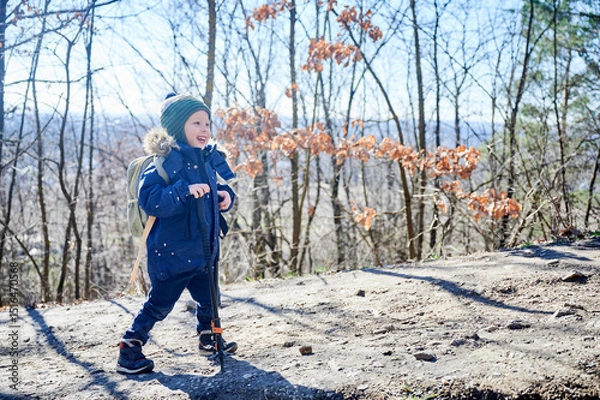 Fototapeta Young child smiling while hiking on forest trail, bundled up in blue winter jacket with fur-lined hood and green striped beanie. Kid holding two trekking poles and enjoying sunny day outdoors.