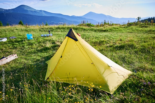 Fototapeta Yellow tourist tent on grassy hillside with camping gear scattered nearby. Stunning view of green hills, distant mountains under clear blue sky, creating perfect spot for peaceful outdoor adventure.
