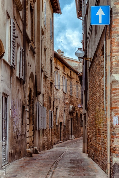Fototapeta A narrow street in Urbino, a medieval town in Italy, where historic buildings with rustic stone facades tower on either side, creating a serene atmosphere.