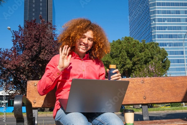 Fototapeta Young man with curly hair waving during a video call, working outdoors with laptop and reusable cup. Concept of remote work, green lifestyle and sustainability