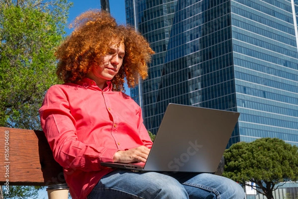 Fototapeta Low angle portrait of a Young man with curly red afro hair working on a laptop in the city, sitting on a bench. Remote work and digital nomad lifestyle concept. Office building on the background