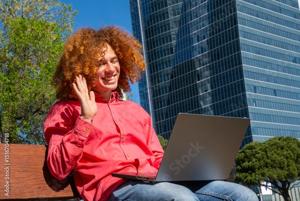 Fototapeta Side view oaf a young man with curly hair waving during a video call, working outdoors with laptop and reusable cup. Concept of remote work, green lifestyle and sustainability