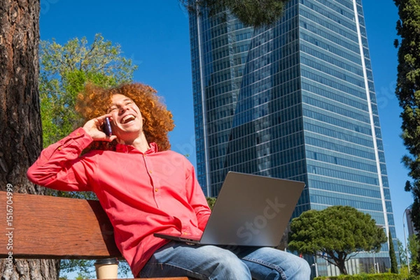 Fototapeta Wide view of a Smiling young man with curly hair smiling and talking on the phone while working on a laptop outdoors in a bussines area. Concept of remote work, communication and sustainable lifestyle