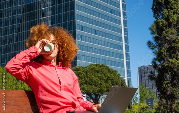 Fototapeta Young man with curly hair drinking from a reusable cup while working on a laptop outdoors. Concept of sustainable habits, remote work and eco-friendly urban lifestyle