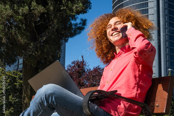 Fototapeta Cheerful young man with red curly hair having a phone call while using a laptop outdoors in a modern business district. Symbol of mobile work, eco-conscious living and digital connection