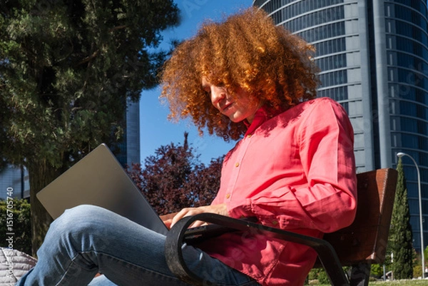 Fototapeta Confident young man with curly red hair sits on a bench working on his laptop in an urban setting. Modern office buildings behind him highlight the digital nomad and remote work lifestyle