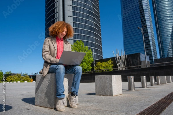 Fototapeta Smiling young man with curly red hair working outdoors on a laptop in front of corporate skyscrapers. Dressed in a casual blazer, he represents modern freelance work and eco-conscious business living