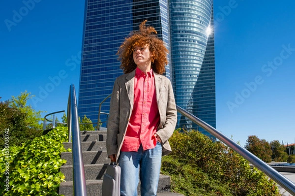 Fototapeta Young man with curly hair walking down stairs in a financial district, holding a laptop case. Modern office tower in background. Concept of green mobility, remote work and urban lifestyle