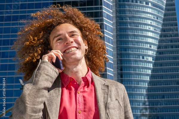 Fototapeta Close-up of a smiling young man with curly hair making a phone call in front of modern office towers. Concept of connection, communication and confident urban professional