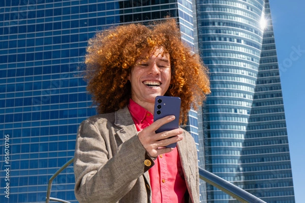 Fototapeta Close-up of a young man with curly hair smiling at his phone in front of a modern office building. Concept of memes, social media, digital connection and contemporary urban lifestyle