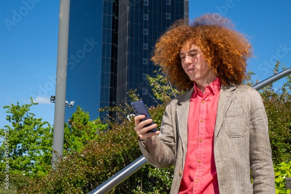 Fototapeta Young man with curly hair focused on his phone while sending a message in a financial district. He wears a blazer, with glass office buildings in the background. Urban freelance lifestyle