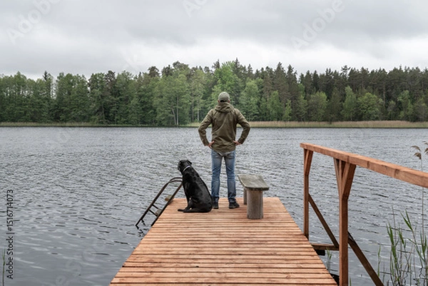 Fototapeta Man and Dog Standing on Wooden Dock Looking Across Forest Lake – Companionship and Reflection in Nature