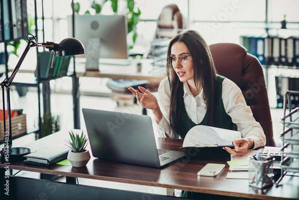 Fototapeta Professional businesswoman in stylish attire working at her desk in an office environment, managing documents and laptop.