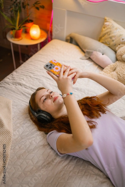Obraz A teenage girl with headphones on her head uses a mobile phone in the bedroom. She's lying on the bed.