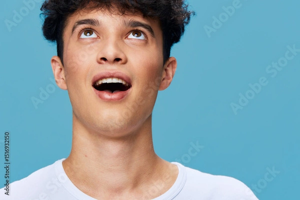 Fototapeta young man with curly hair looking up with surprised expression on blue background wearing white shirt, capturing emotion and youth vitality