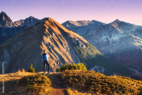Obraz Young woman with backpack standing on grassy mountain trail in valley with high mountain peaks during golden hour sunset in autumn. Hiking and trekking in Italy, Livigno. Girl in Alps in fall