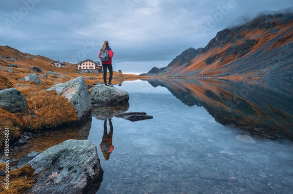 Obraz Young woman hiker with backpack on a rock in autumn. Girl and breathtaking view of a mountain alpine lake reflecting the surrounding peaks, orange grass and cloudy dramatic sky. Switzerland in fall